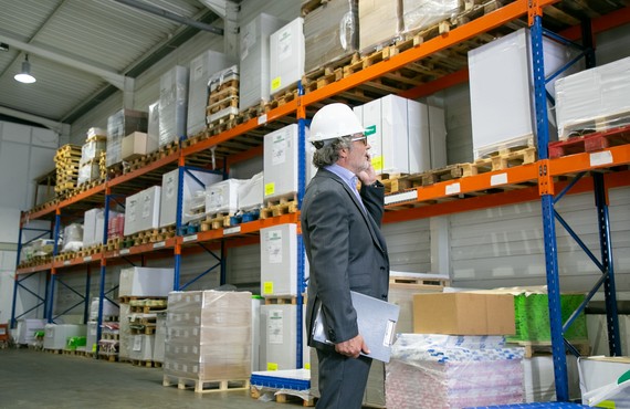 Focused CEO in formal suit and hardhat standing in warehouse and talking on mobile phone. Shelves with goods on wooden pallets. Side view. Business or logistics concept